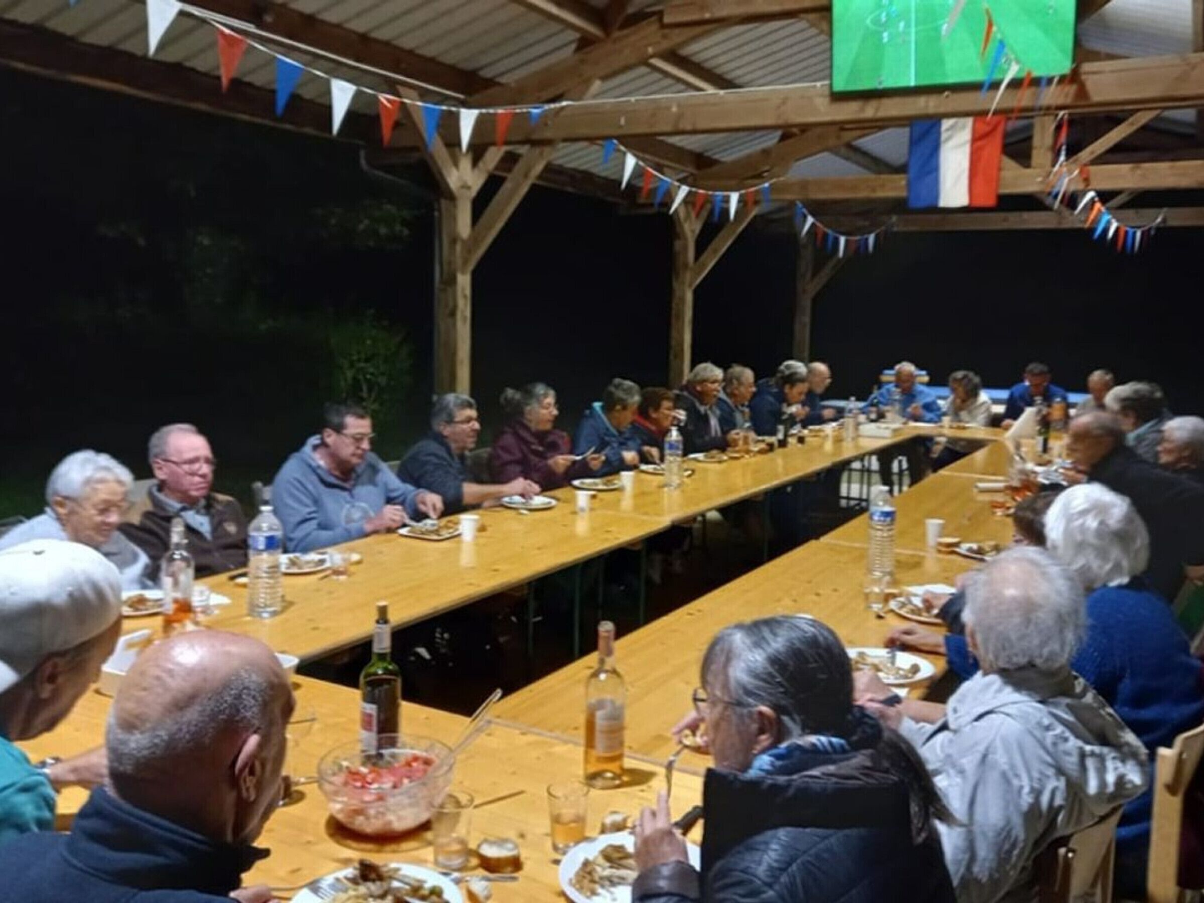 A long wooden table with people seated around it, under a covered area with a roof.