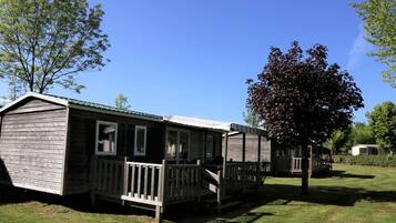 A single-story mobile home with a porch and a small deck, surrounded by grass and trees.