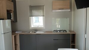 A compact kitchen with a sink, stove, and cabinets, a window with blinds, and a white chair. - Cozy mobile home - 2 guests (Le Château-d'Oléron)