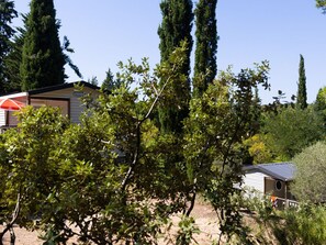 A house with a visible garage door, surrounded by dense foliage and tall trees. - Mobile-home with AC, pets welcome - 4 guests (Lézignan-Corbières)