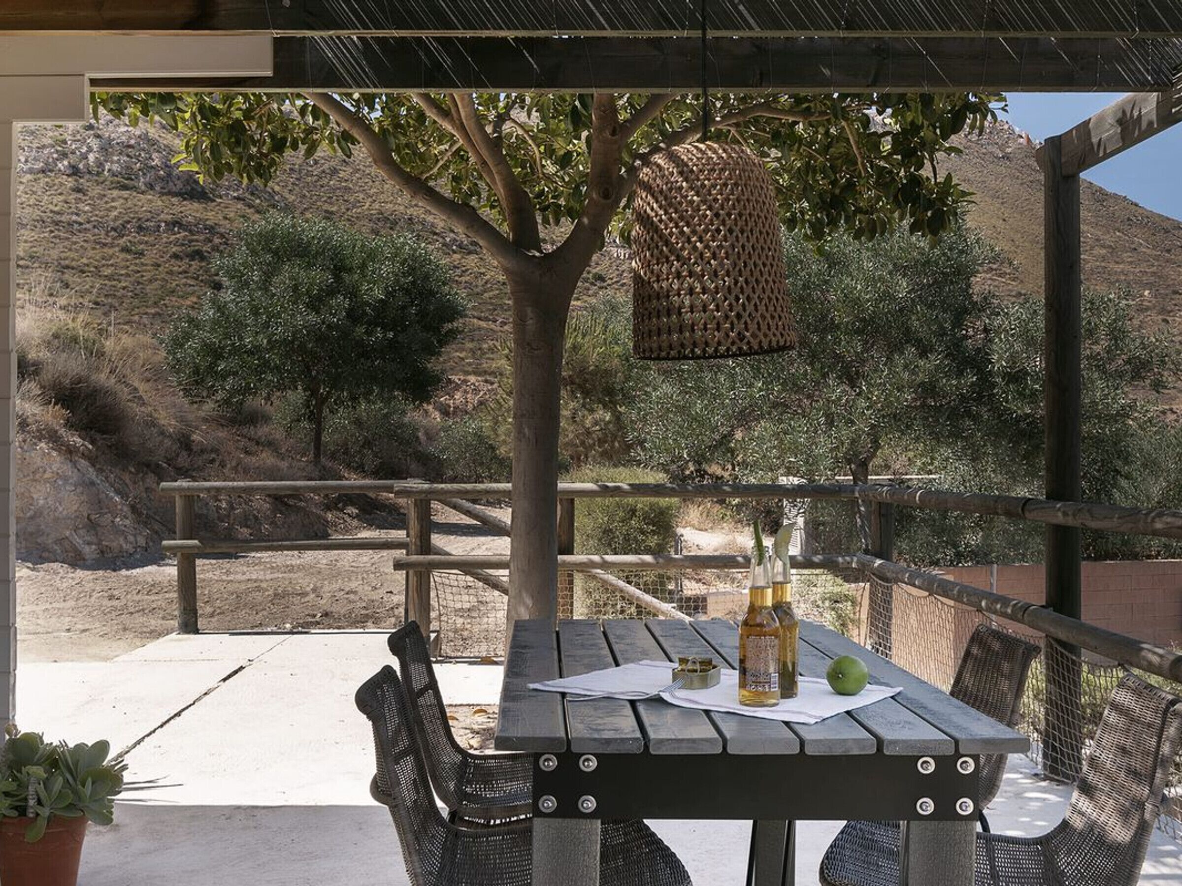 An outdoor patio with a wooden table and chairs, a hanging basket, and a view of a hilly landscape.