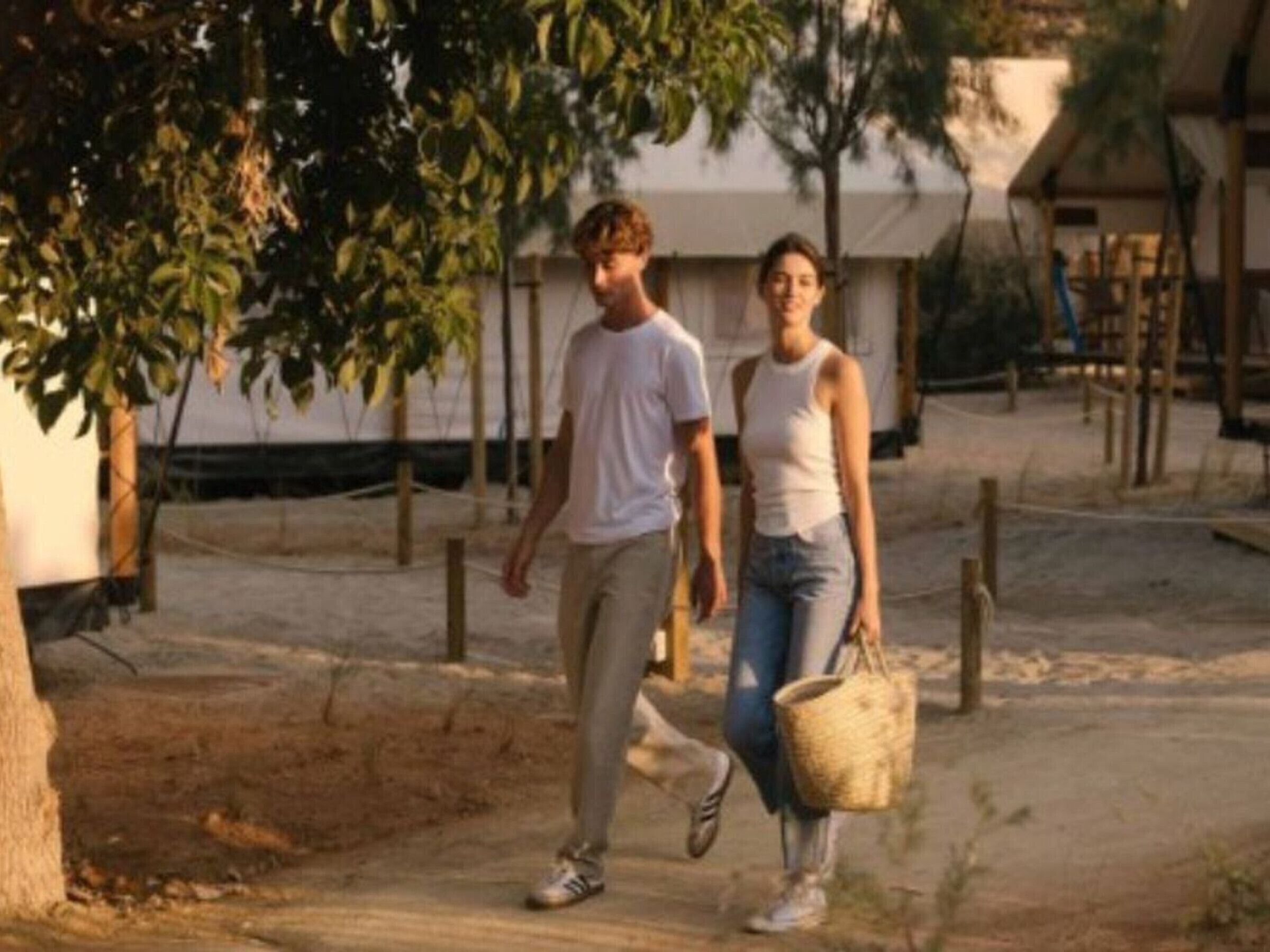 Two people walking on a dirt path with trees and a building in the background.