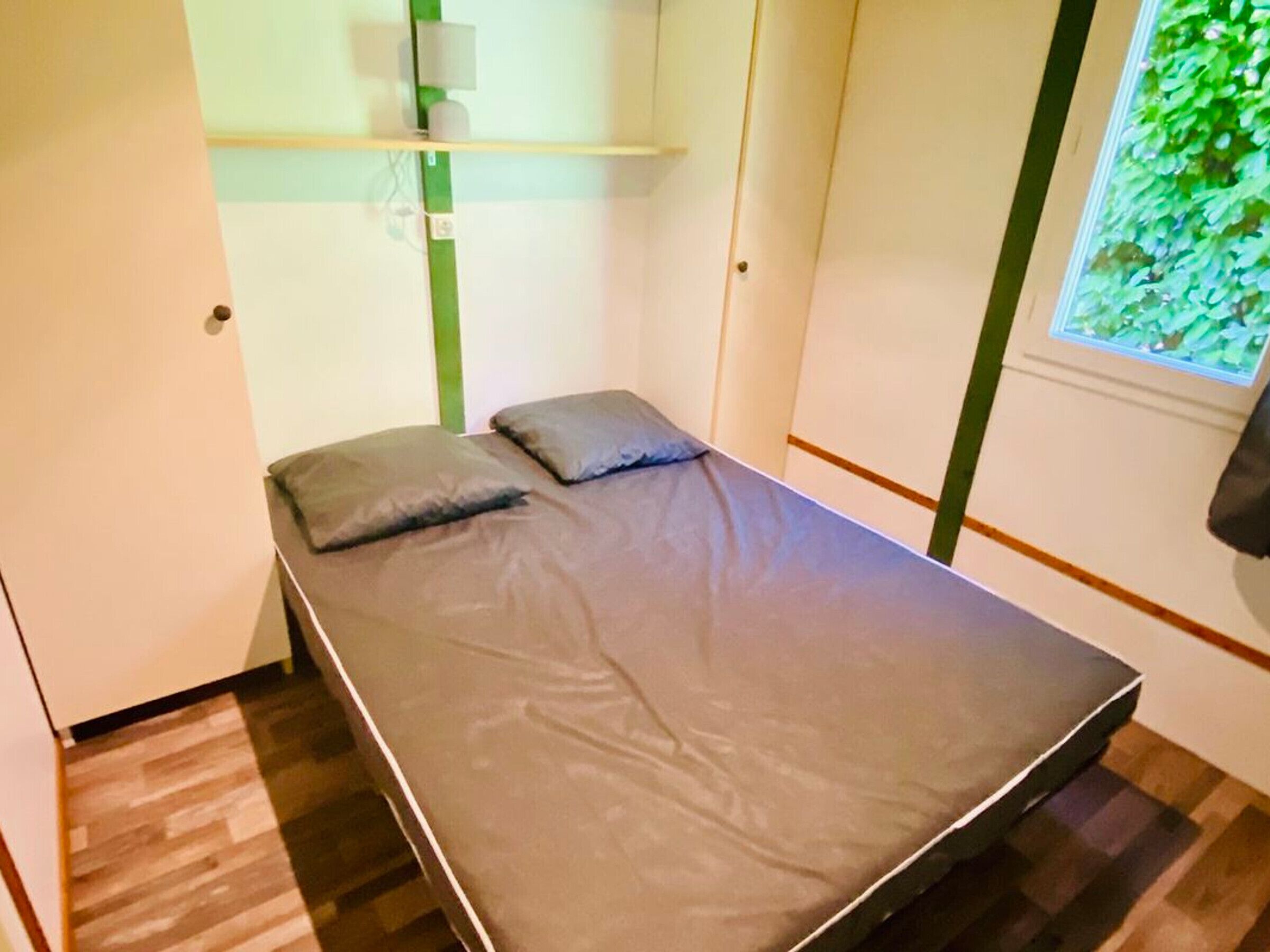 A small, single-bed room with a grey mattress, two pillows, a wooden floor, a window with a green plant view, and a white wall with a shelf above the bed.