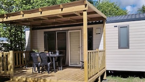 A covered outdoor area with a table and chairs, a wooden deck, and a sliding glass door leading to a house. - Mobile home, pets welcome - 4 guests (Chaillé-les-Marais)
