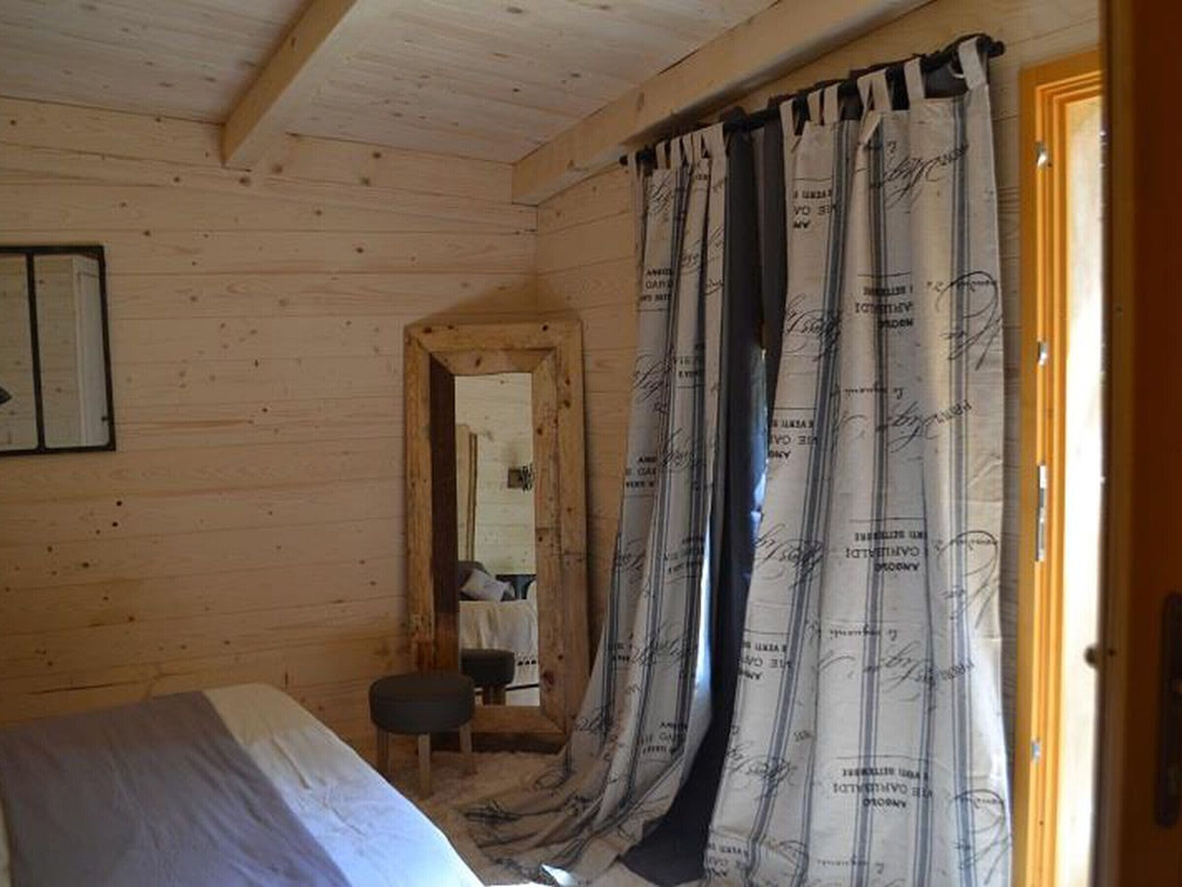 Una habitación de cabaña de madera con cama, un taburete pequeño, un espejo y una puerta.