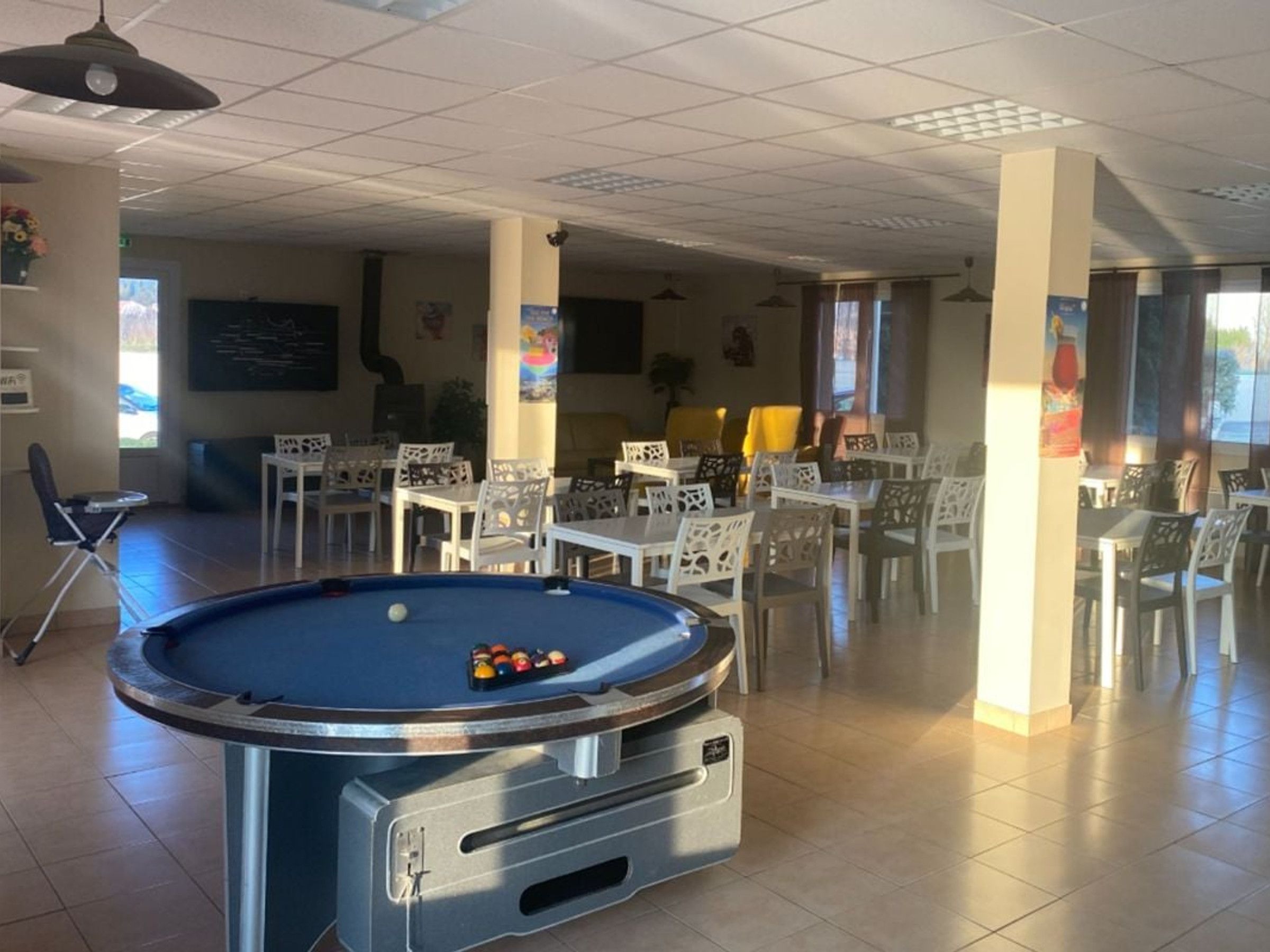 A pool table with balls, surrounded by white chairs and tables in a room with tiled floors and ceiling lights.