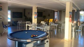 A pool table with balls, surrounded by white chairs and tables in a room with tiled floors and ceiling lights.