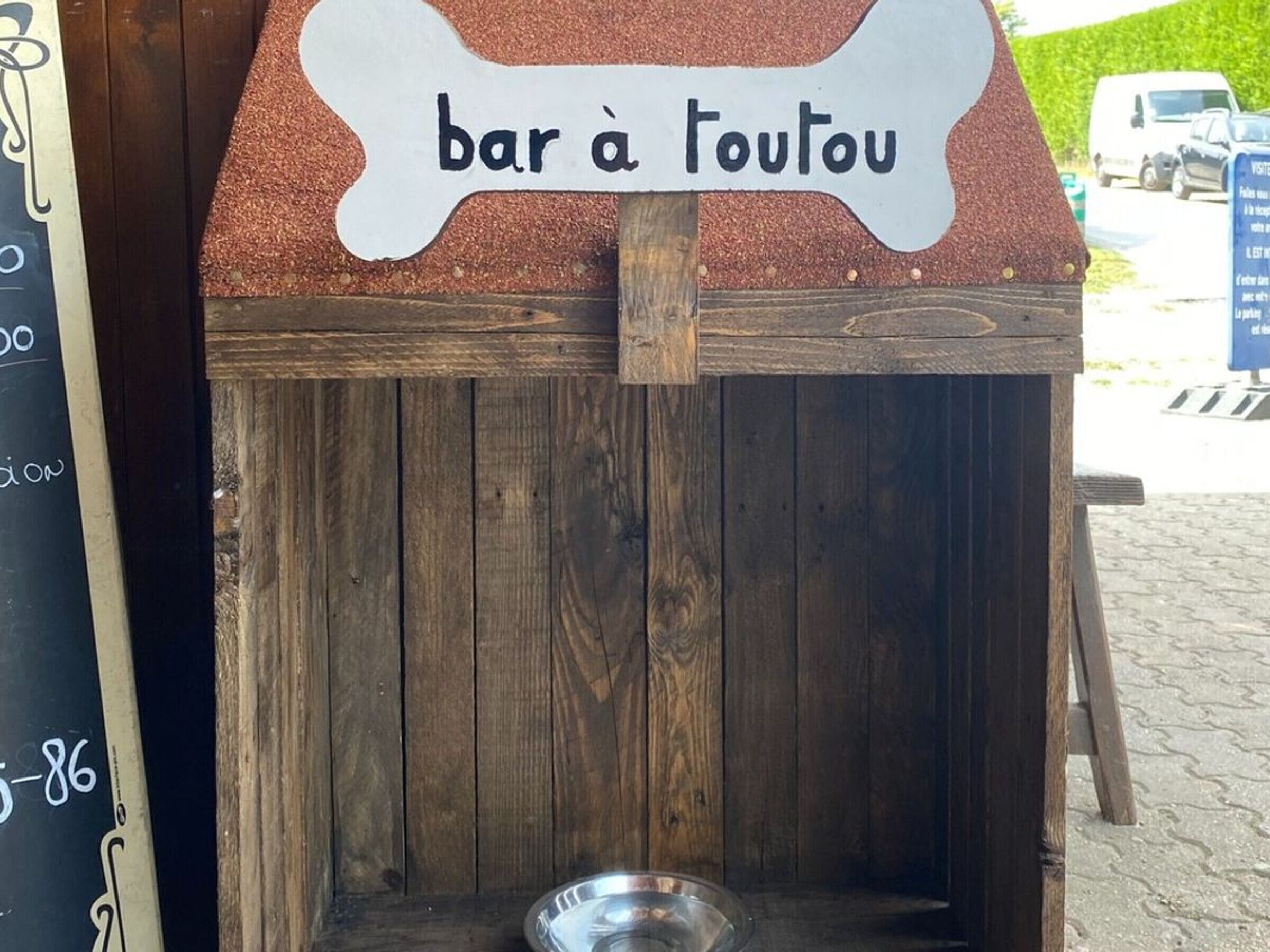 En rustik träkonstruktion med en skylt där det står ”bar à toutou” och en metallskål på marken.