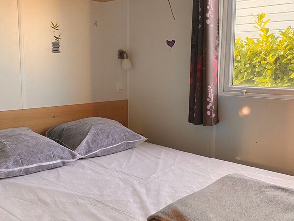 A bed with pillows, a bedside lamp, a wall decoration, and a window with curtains. - Cozy mobile home with AC – 2 guests (Angicourt)
