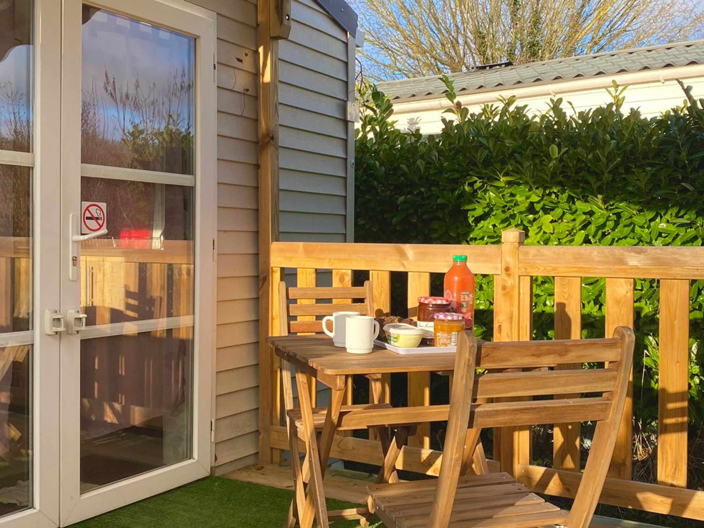 Een houten terras met een tafel en stoelen, een glazen deur met een rookverbods bordje, en een groene haag op de achtergrond.