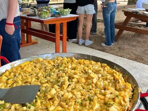 Una sartén metálica grande llena de una preparación de papas y queso, sobre una mesa de madera al aire libre.