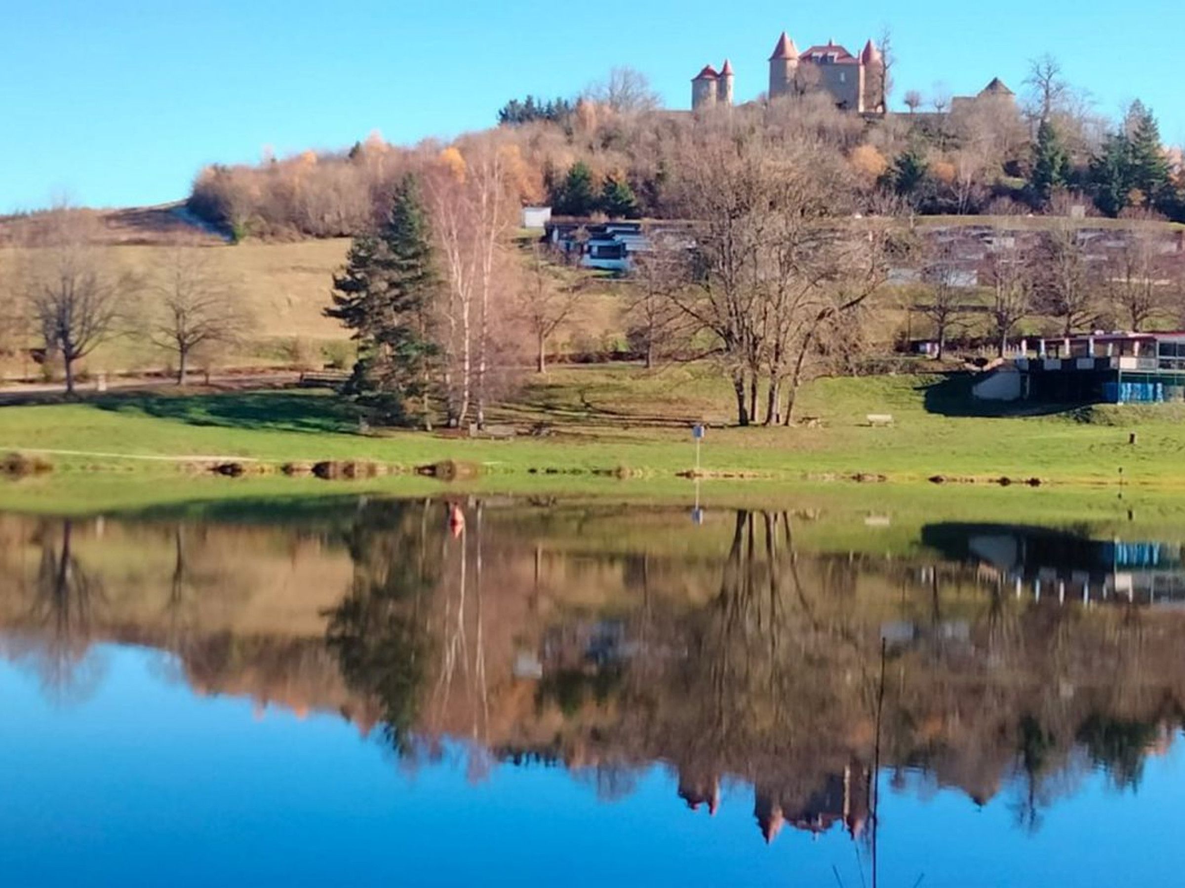 A calm lake with a clear reflection, surrounded by grassy areas and trees. In the background, there is a castle-like structure on a hill.
