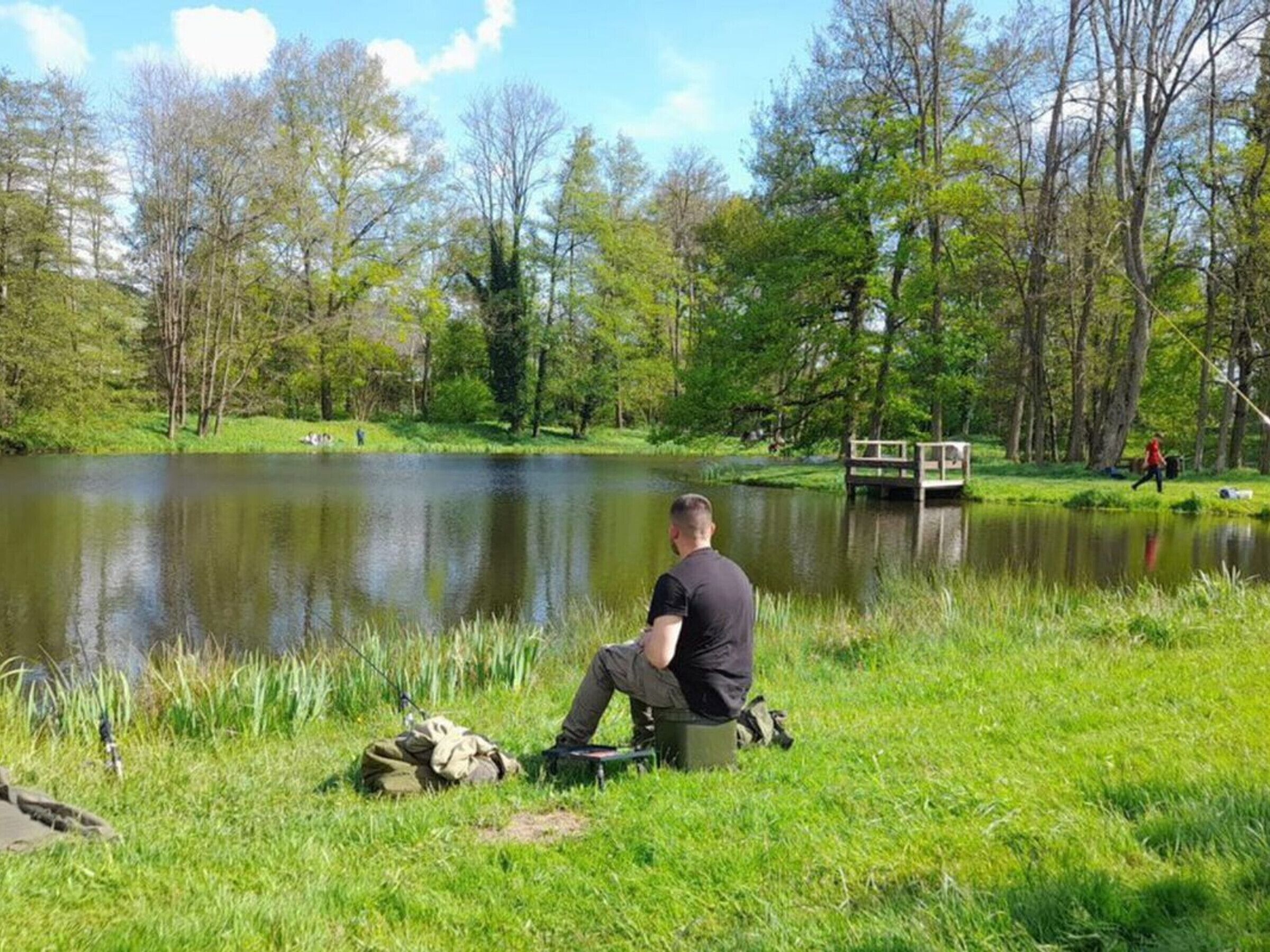 Mies istuu järven rannalla, taustalla näkyy puita ja puinen silta.
