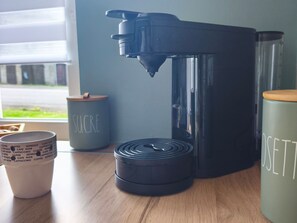 Une machine à café, une tasse ornée d’un motif ou d’une inscription sur le thème du café, et deux récipients munis de couvercles, le tout posé sur une table en bois.