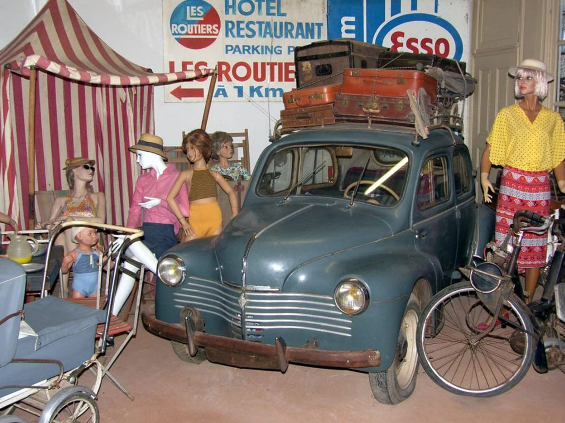 A vintage car with luggage on the roof, a wheelchair, and mannequins in the foreground.