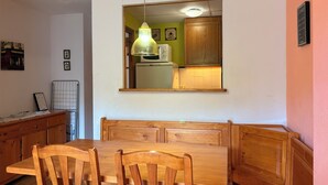 A dining area with a wooden table and chairs, a hanging light fixture, and a view into a kitchen with a refrigerator and wooden cabinets. - Mountain environment. Fireplace and garden - Isard 4 (Pla de l'Ermita)