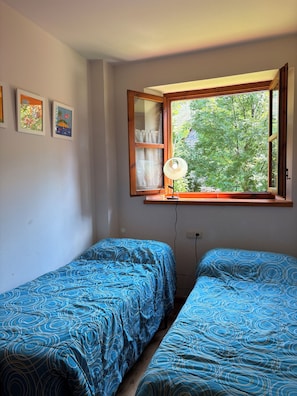 Une chambre avec deux lits simples, une fenêtre donnant sur un espace verdoyant et une étagère murale avec une lampe.