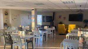 A dining area with white tables and chairs, a yellow sofa, and a TV mounted on the wall.