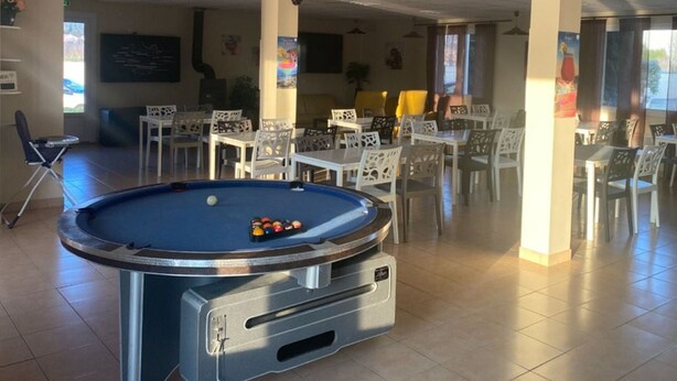 Una mesa de billar con bolas, rodeada de sillas y mesas blancas en una habitación con piso de baldosas e iluminación en el techo.