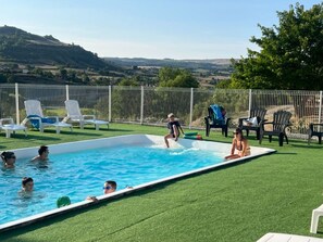 A swimming pool with people in the water and on the edge, surrounded by lawn chairs and a white fence.