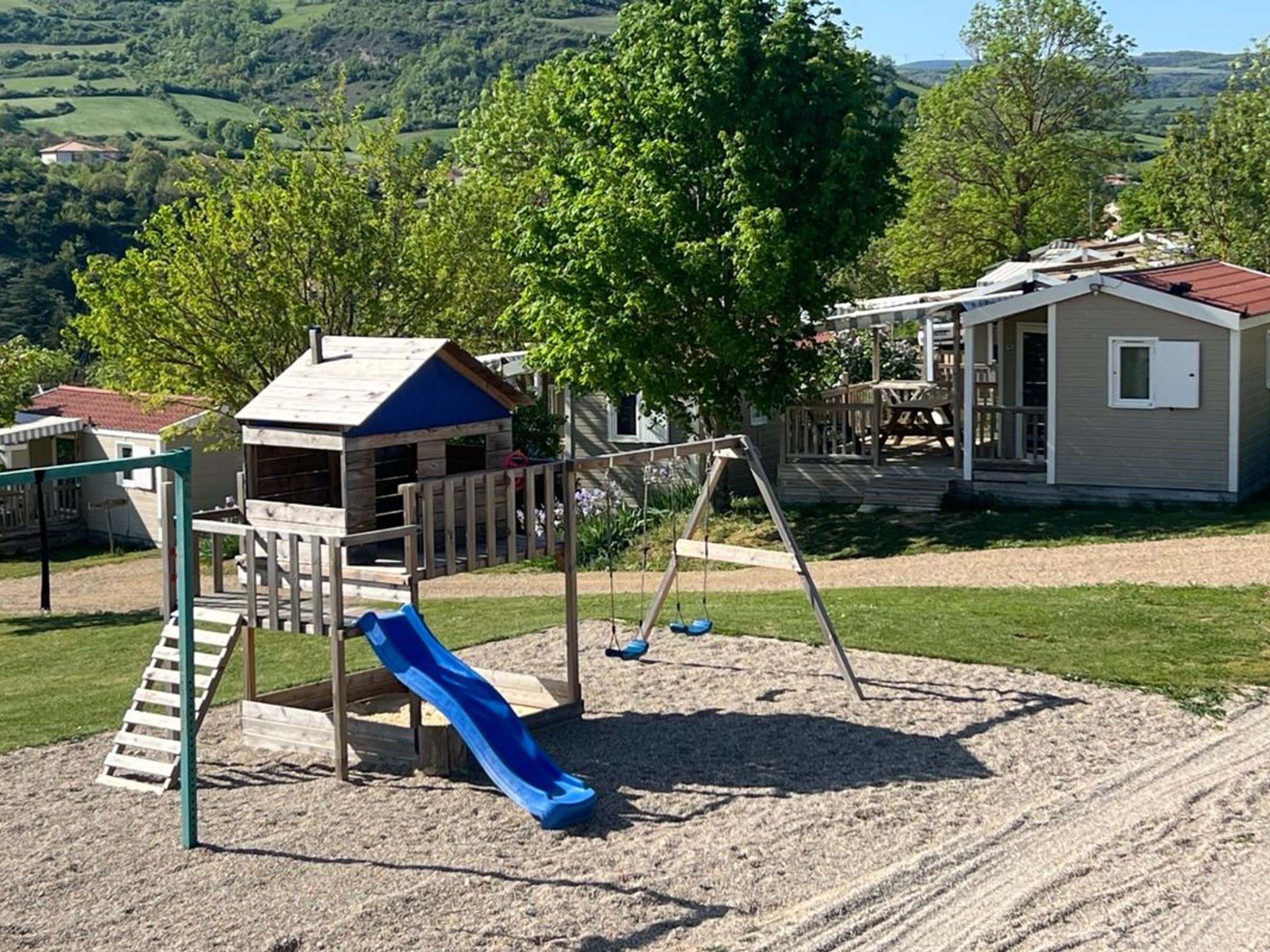 Un parque infantil con tobogán y columpios, rodeado de césped y un edificio con techo rojo.