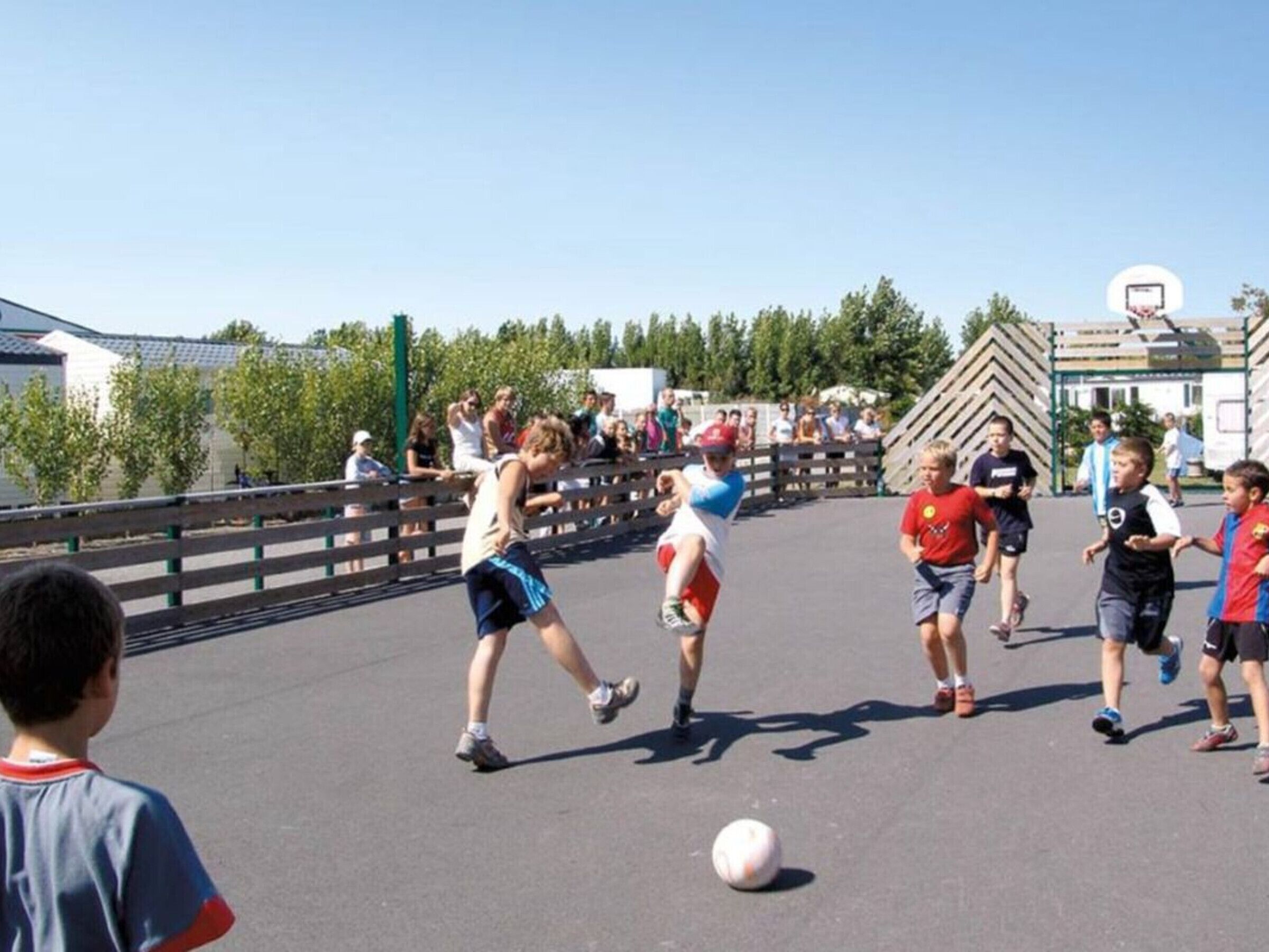 Un grupo de niños jugando al fútbol en un área pavimentada con una valla y árboles al fondo.