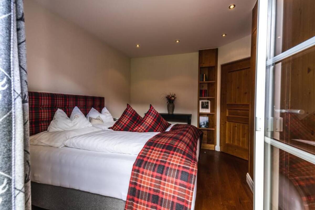 A bedroom with a bed, a red plaid blanket, a wooden headboard, a bookshelf, and a window.