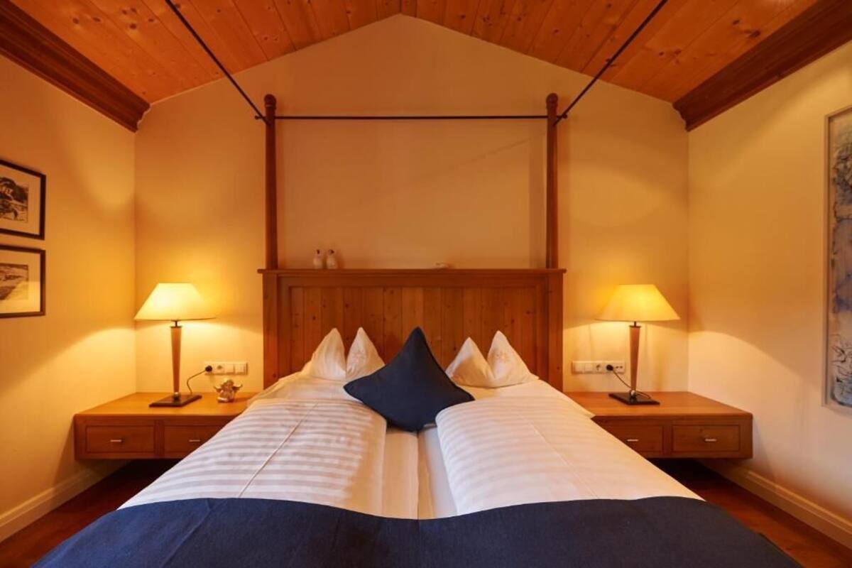 A bedroom with a wooden ceiling, a bed with white bedding and two pillows, two bedside tables with lamps, and a painting on the wall.
