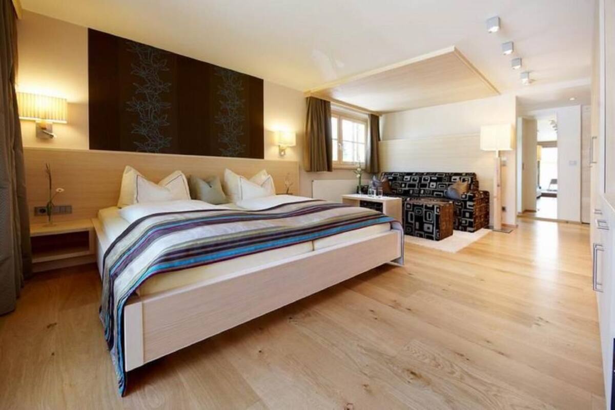 A modern hotel room with a large bed, a desk, and a seating area with patterned cushions.