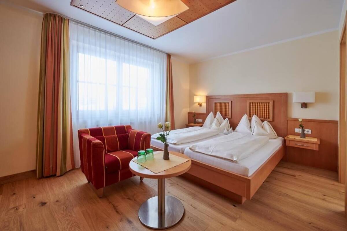 A hotel room with a bed, a red armchair, a small round table, and a window with sheer curtains.