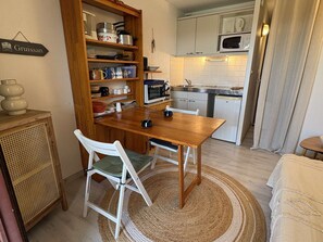 A compact kitchen with wooden cabinets, a microwave, a stove, and a dining area with a table and chairs. - Loft studio, AC & pool - 2 adults, 2 kids (Gruissan)