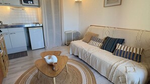 A compact living space with a kitchenette, a sofa with patterned cushions, a small wooden table, and a woven rug. - Loft studio, AC & pool - 2 adults, 2 kids (Gruissan)