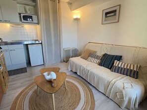 A compact living space with a kitchenette, a sofa with patterned cushions, a small wooden table, and a woven rug. - Loft studio, AC & pool - 2 adults, 2 kids (Gruissan)