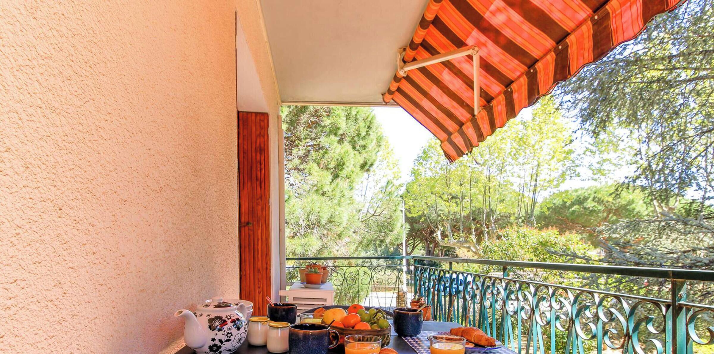 A balcony with a view of trees, a table with breakfast items, and a red awning.