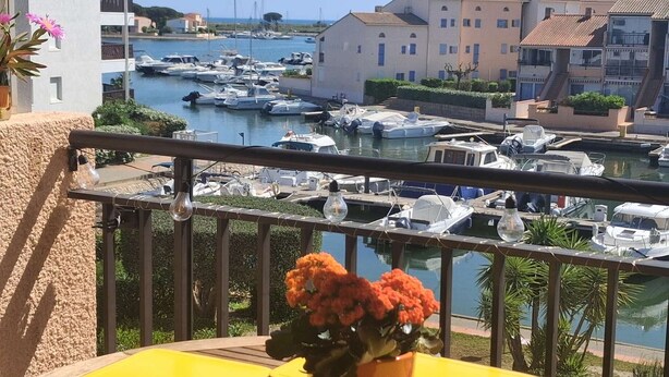 Un balcón con vista a un puerto deportivo, macetas con flores y una mesa amarilla.