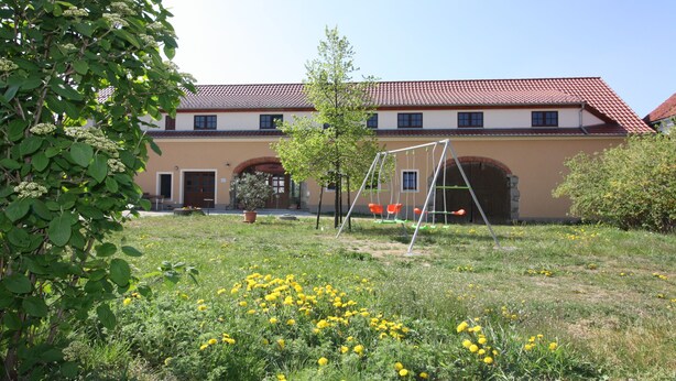 A building with a red-tiled roof, arched doorways, and a grassy area with yellow flowers and a swing set. - Ferienhof Steindörfel (Hochkirch)