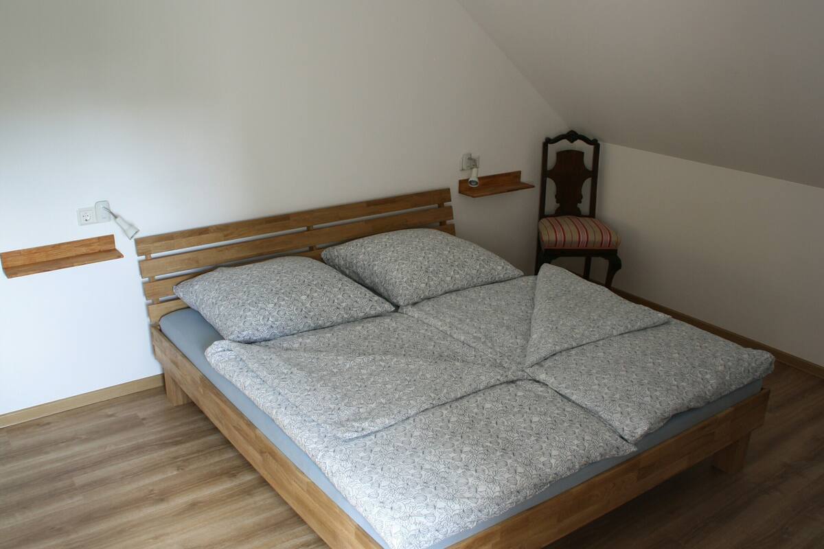 Een enkel bed met houten frame en een beddengoed met dessin, een klein nachtkastje, een stoel met gestreept kussen, en een wandplank.