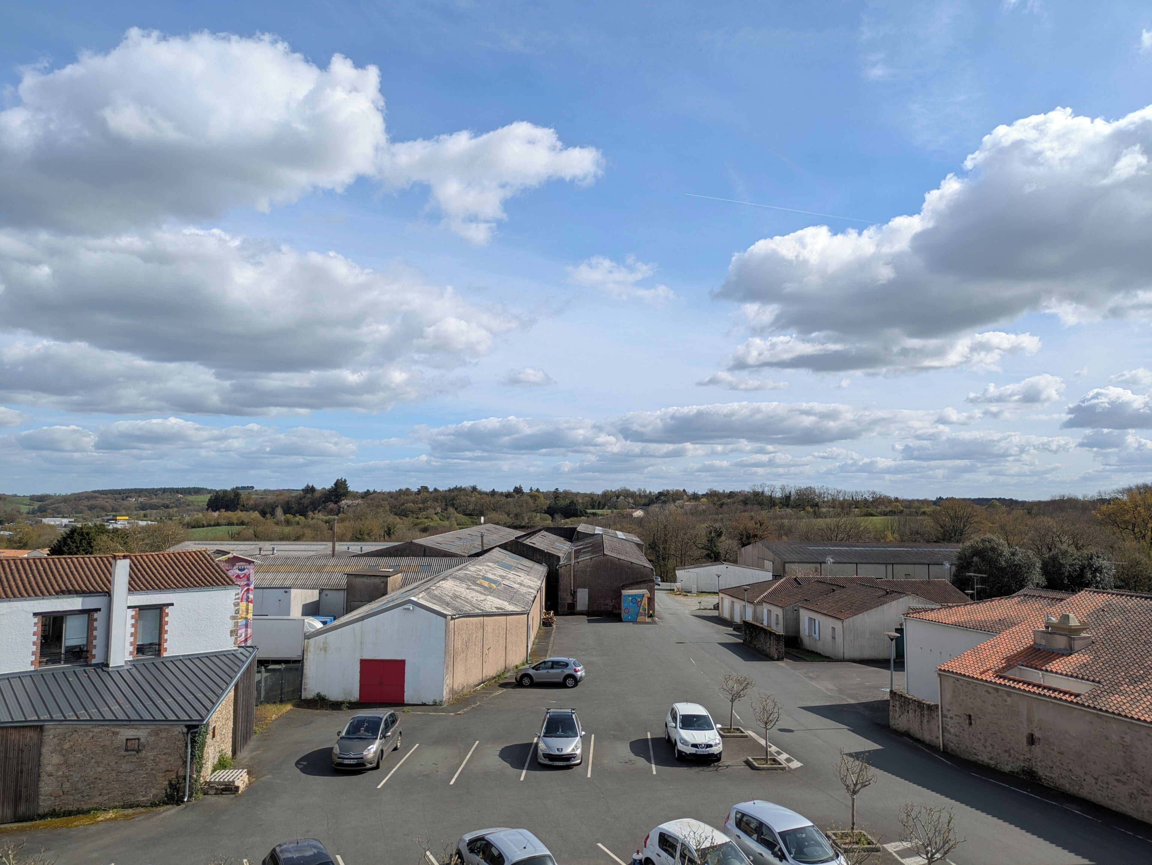 Ein Parkplatz mit mehreren Autos, umgeben von Gebäuden mit roten Ziegeldächern und einem klaren Himmel mit Wolken.