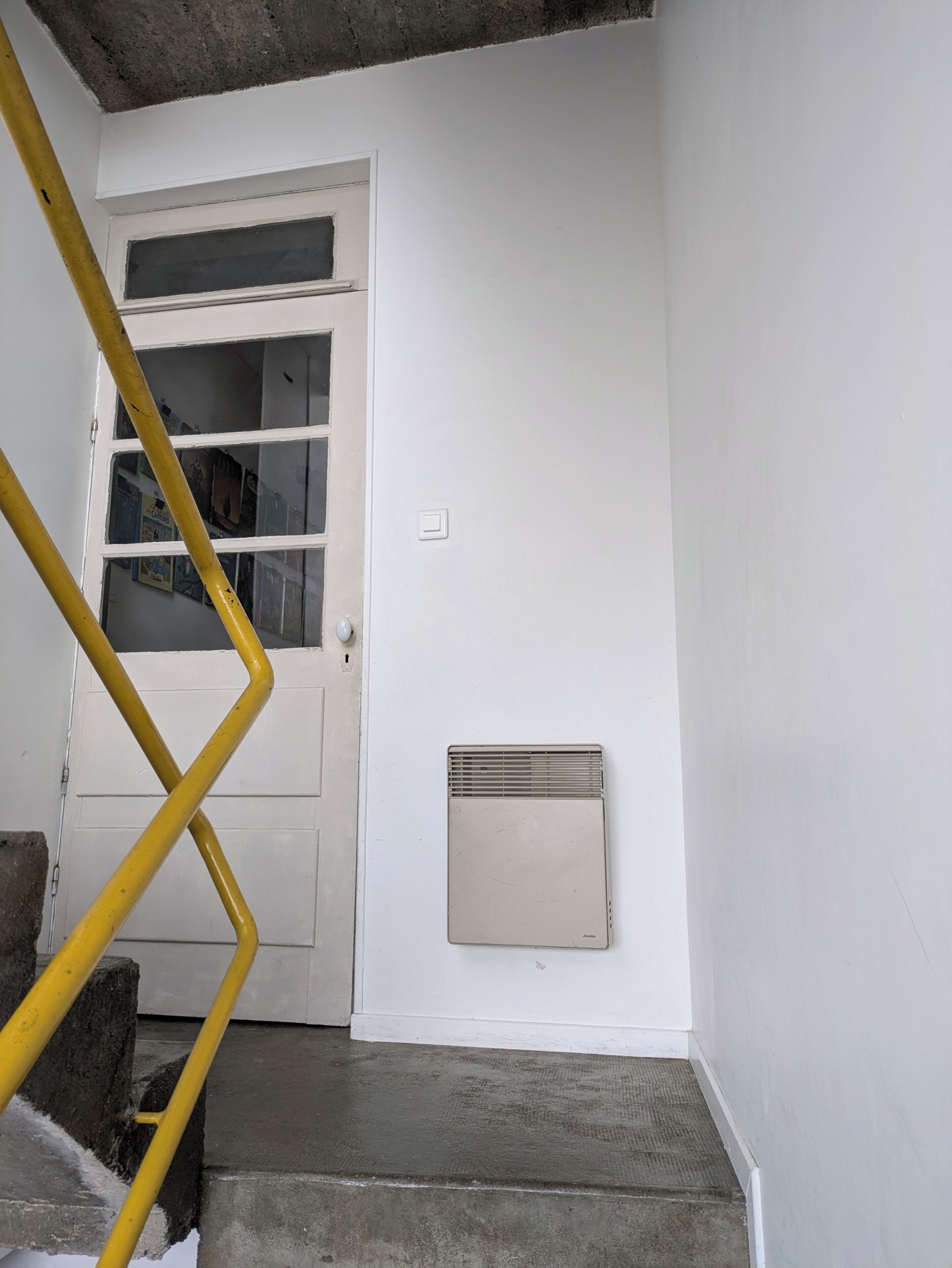 Une porte blanche avec une fenêtre, une rambarde jaune et une bouche d’aération fixée au mur.