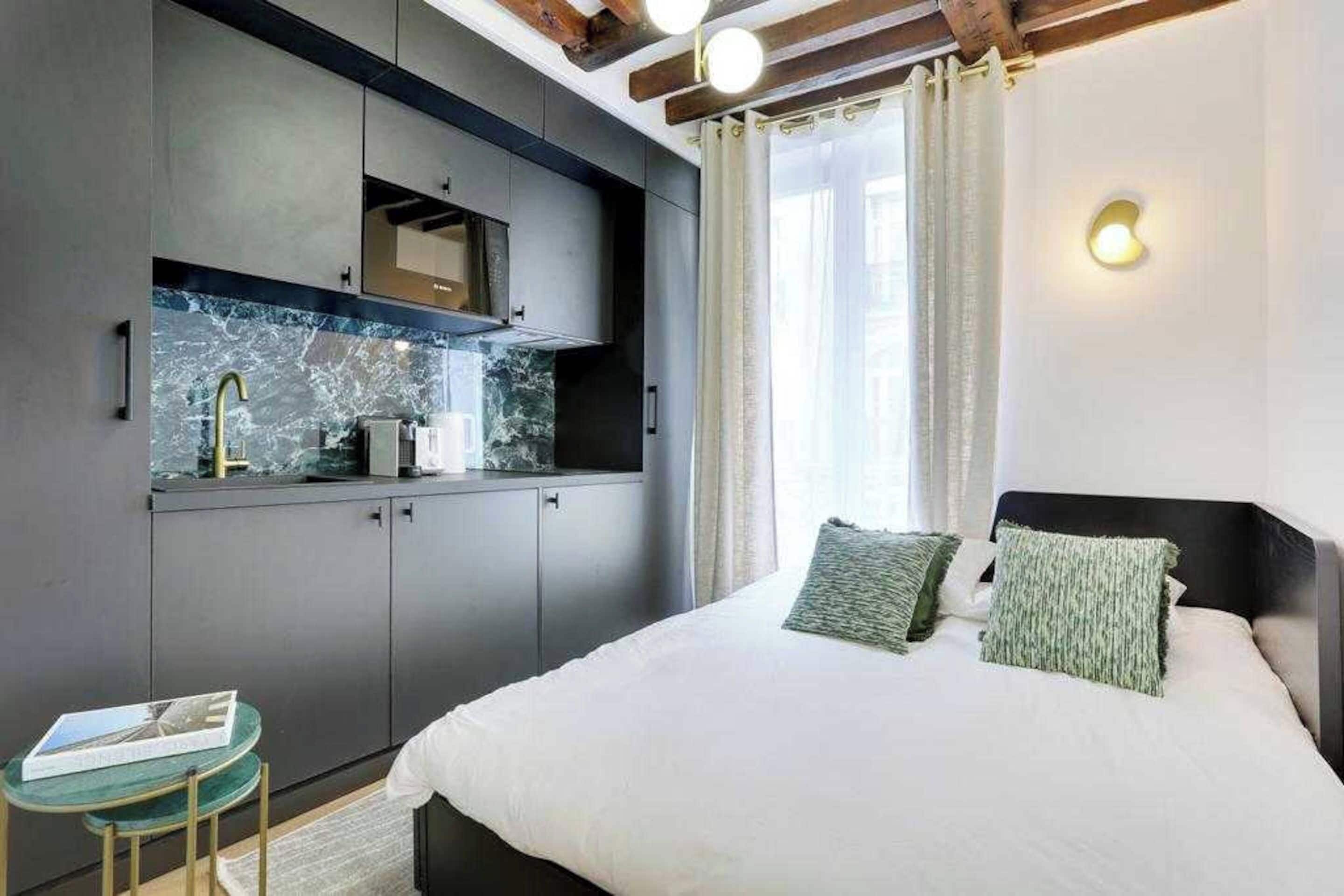 Una camera da letto con un letto, due cuscini, un comodino, un tavolino e un angolo cottura integrato con lavello e pensili.