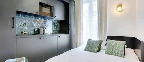 A bedroom with a bed, two pillows, a nightstand, a small table, and a built-in kitchen area with a sink and cabinets.