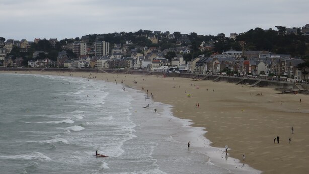 Un pueblo costero con una playa de arena, olas y edificios al fondo.