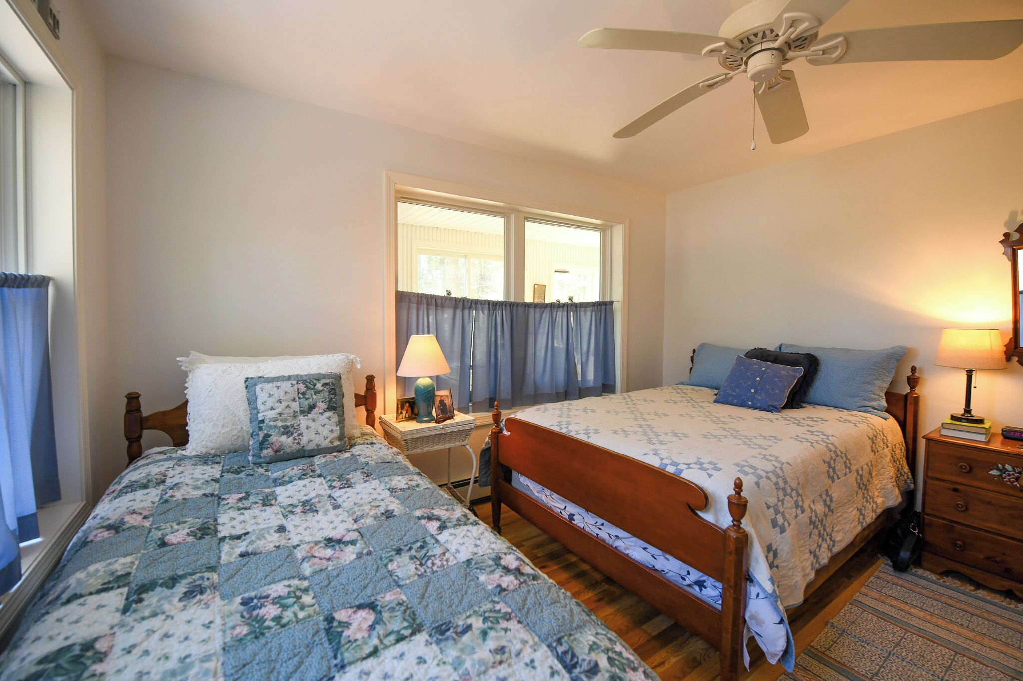 A bedroom with two beds, a ceiling fan, a window with curtains, and a nightstand with a lamp.