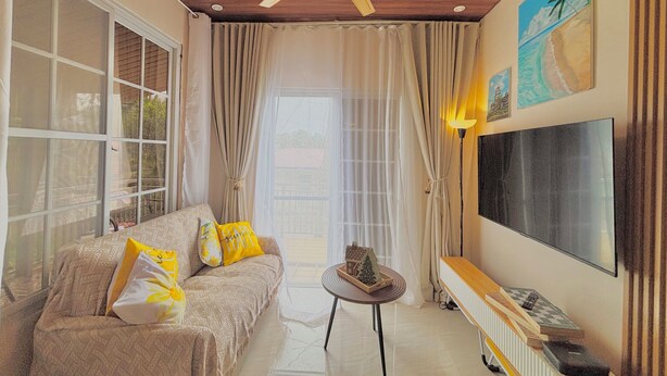 Una sala de estar con un ventilador de techo, un sofá con cojines estampados, una mesita con una casa de modelo, un televisor montado en la pared y una ventana con cortinas.