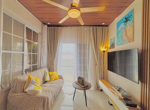 A living room with a ceiling fan, a sofa with patterned cushions, a small table with a model house, a TV mounted on the wall, and a window with curtains.