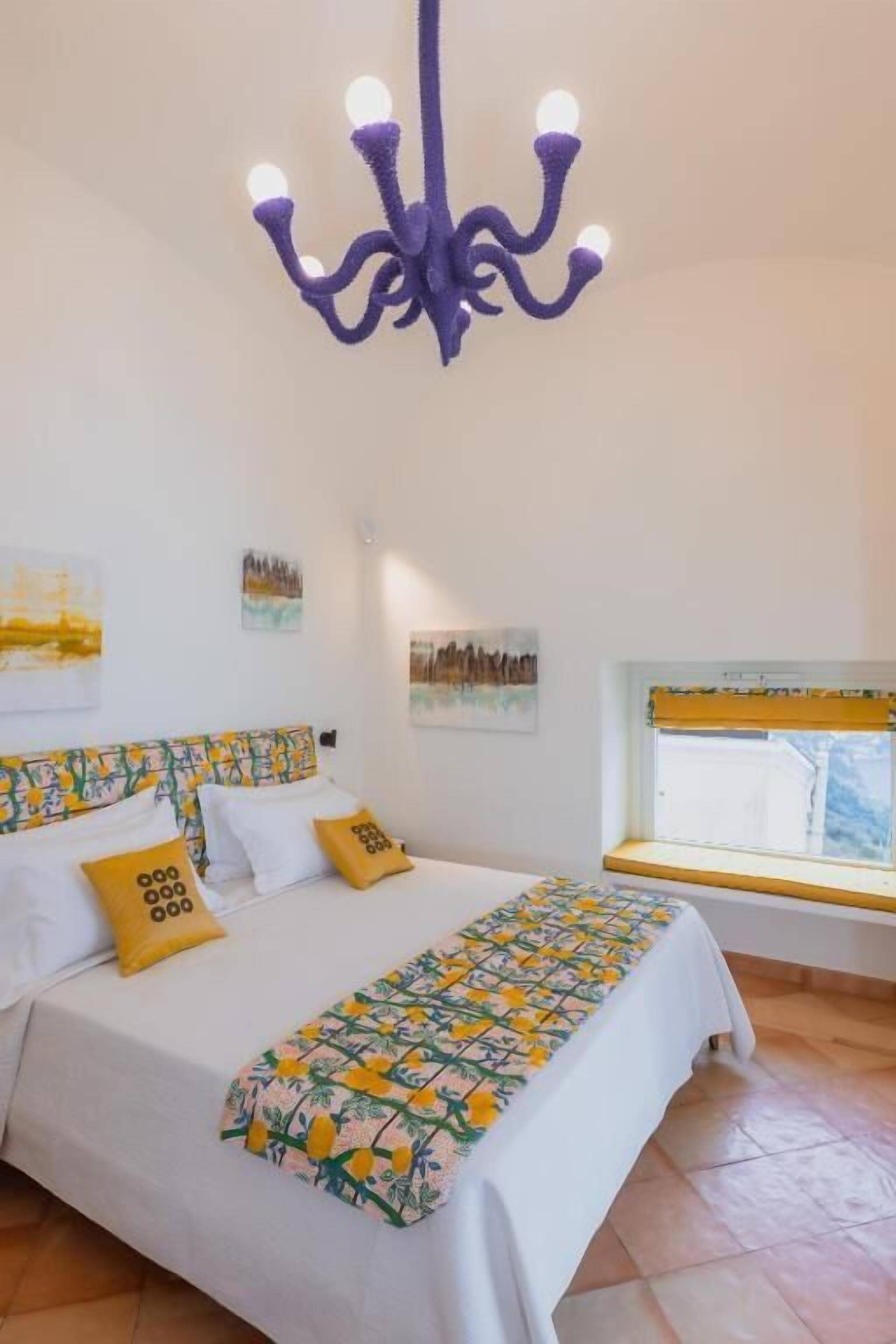 A bedroom with a bed, a window with yellow blinds, a chandelier, and artwork on the walls.