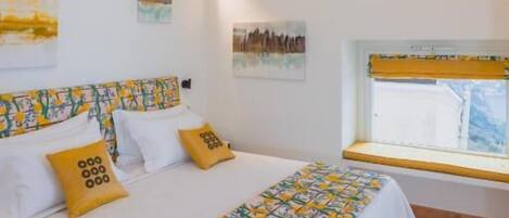 A bedroom with a bed, a window with yellow blinds, a chandelier, and artwork on the walls.