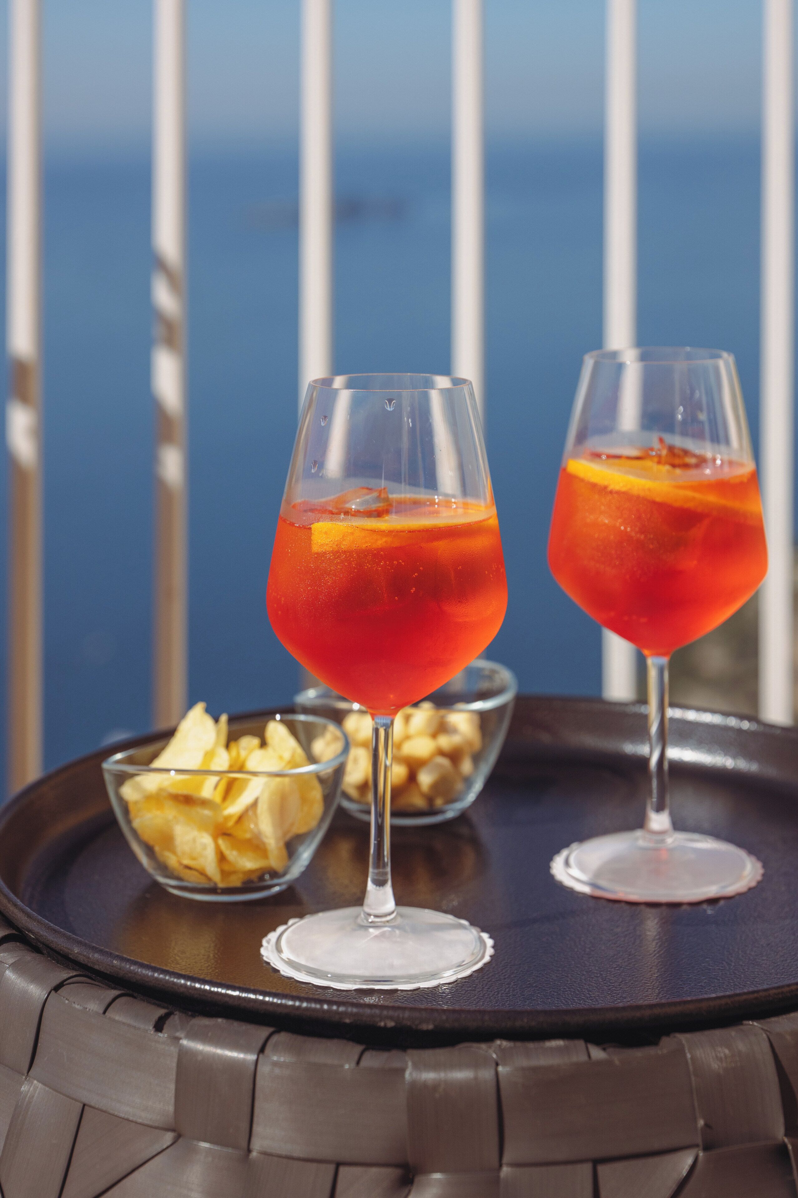 Deux verres d’une boisson rouge, décorés de tranches d’orange, sont posés sur un plateau, à côté d’un bol rempli d’en-cas, et on aperçoit la mer au loin.