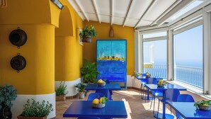 Un espacio de comedor al aire libre iluminado por el sol, con paredes amarillas, mesas y sillas azules, plantas en macetas y vistas al mar.