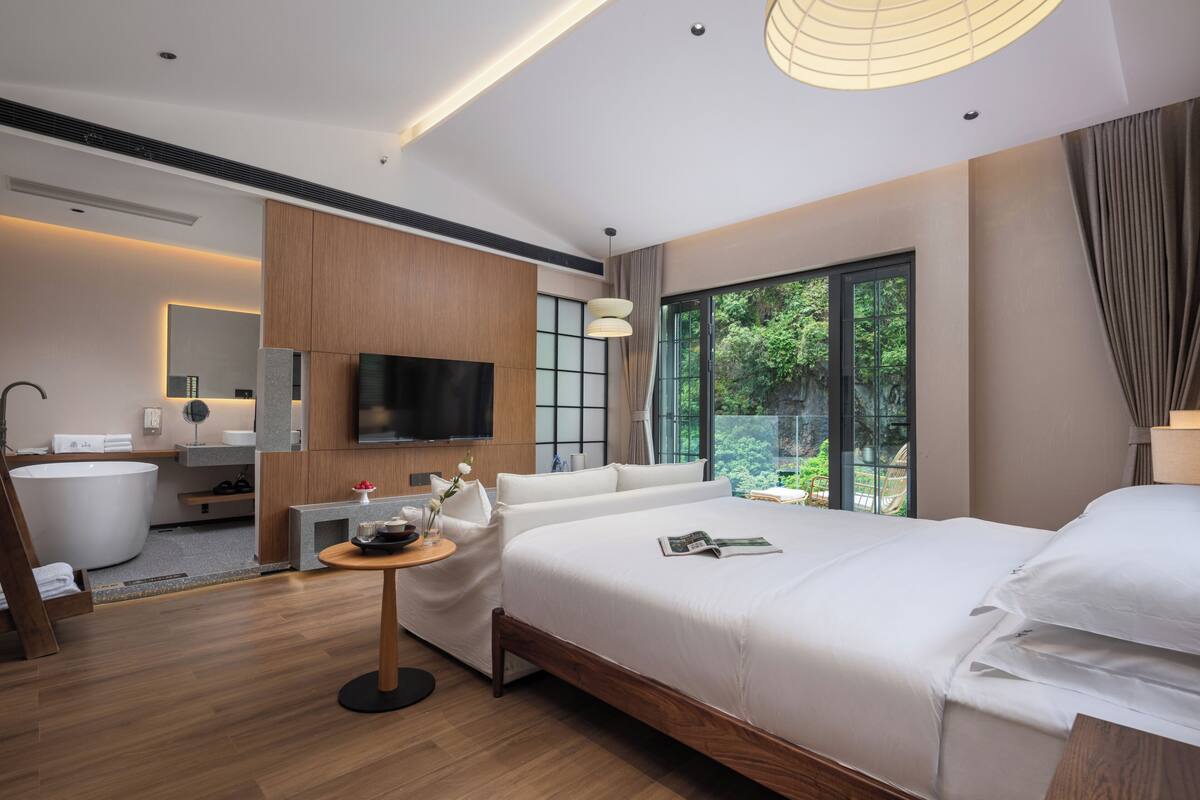 Een moderne hotelkamer met een groot bed, een flatscreen televisie, een badkuip en een groot raam met uitzicht op groen.